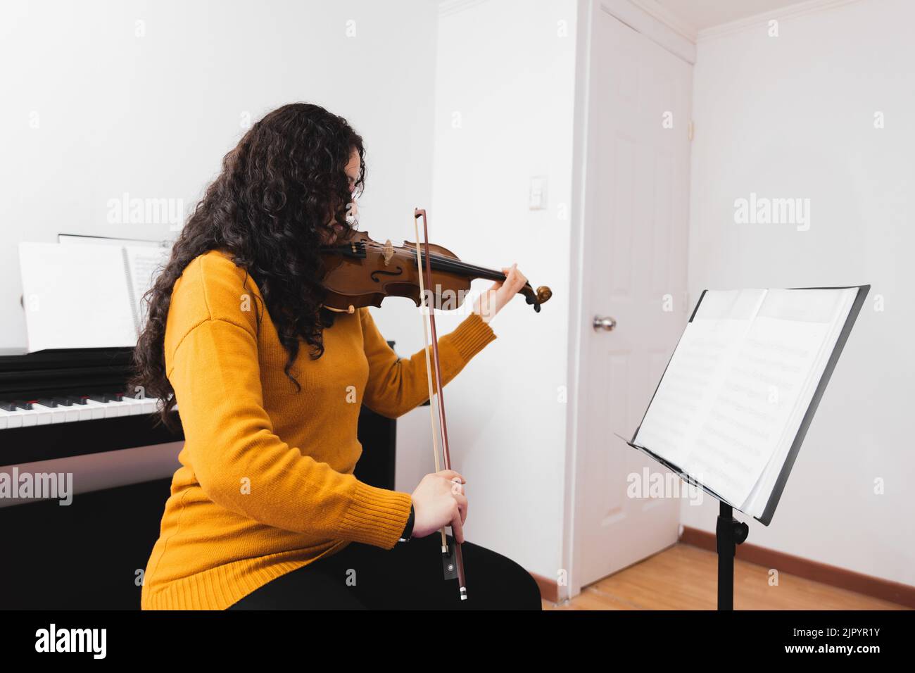 Donna bruna che indossa un maglione giallo e suona violino leggendo spartiti musicali. Foto Stock
