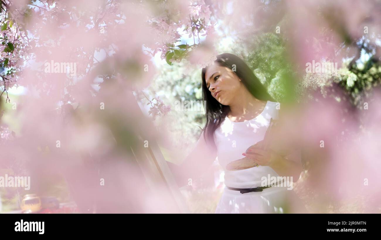 una bella donna pittore in vestito bianco, l'artista dipinge un quadro di fiori in primavera fiorente frutteto di mele, tiene una tavolozza con pitture e un pennello nelle sue mani. Foto di alta qualità Foto Stock