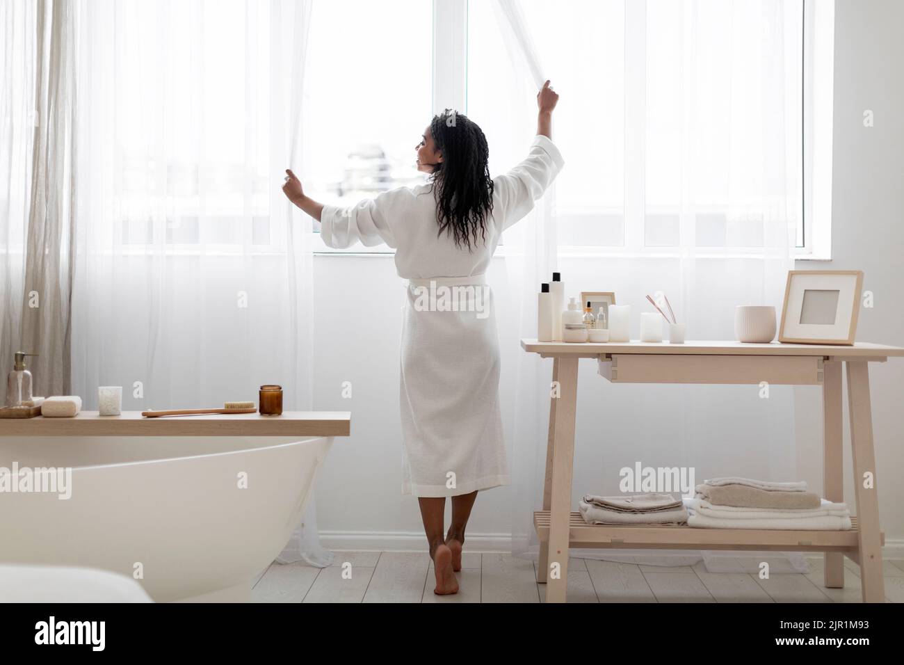 Giovane donna nera che apre le tende mentre si fa mattina di bellezza routine in bagno Foto Stock