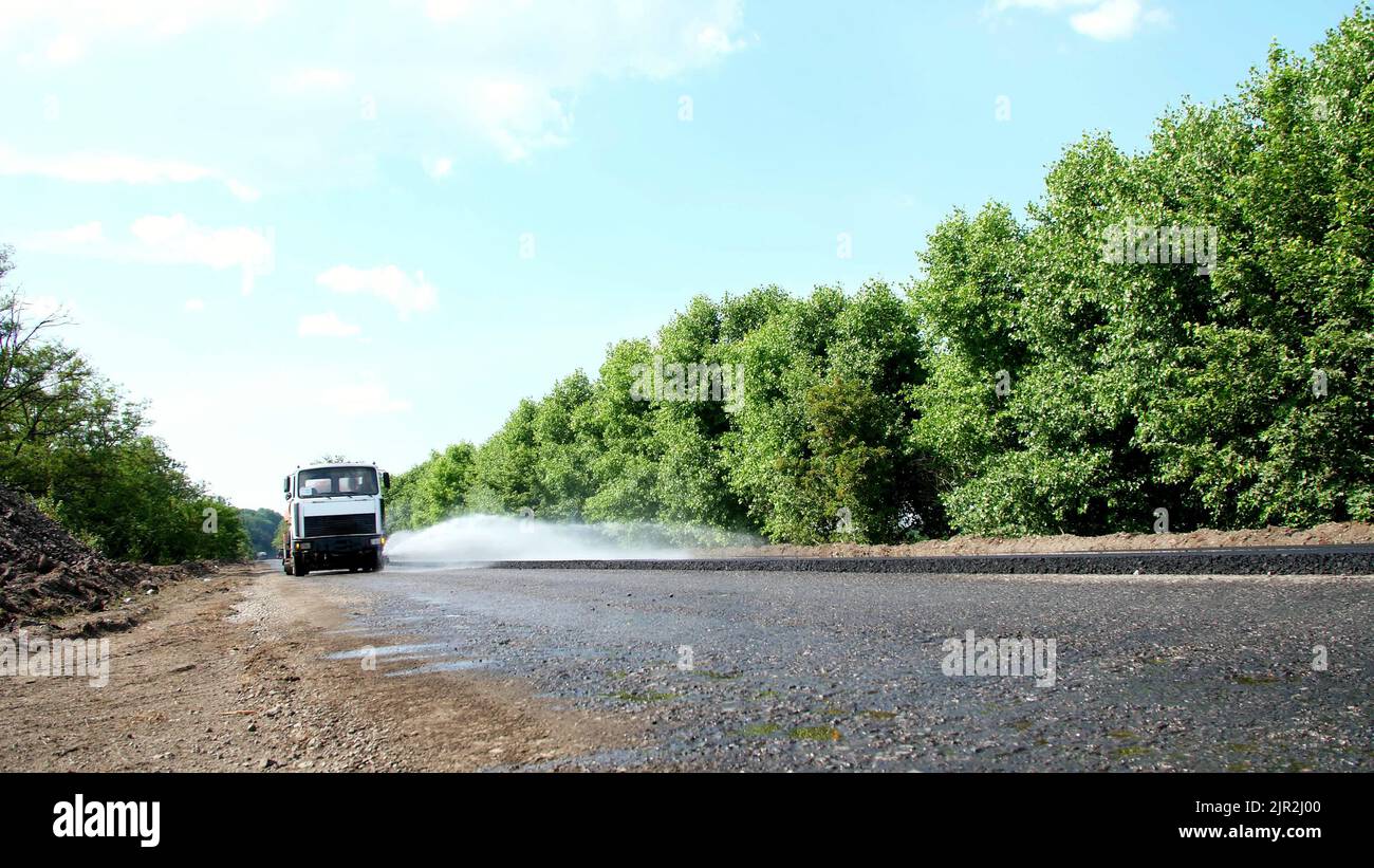 Riparazione di un'autostrada, lavori di costruzione stradale. Una speciale macchina irrigatrice spruzza acqua su una nuova pavimentazione in asfalto fresco, coprendo un lato del traffico. Foto Stock
