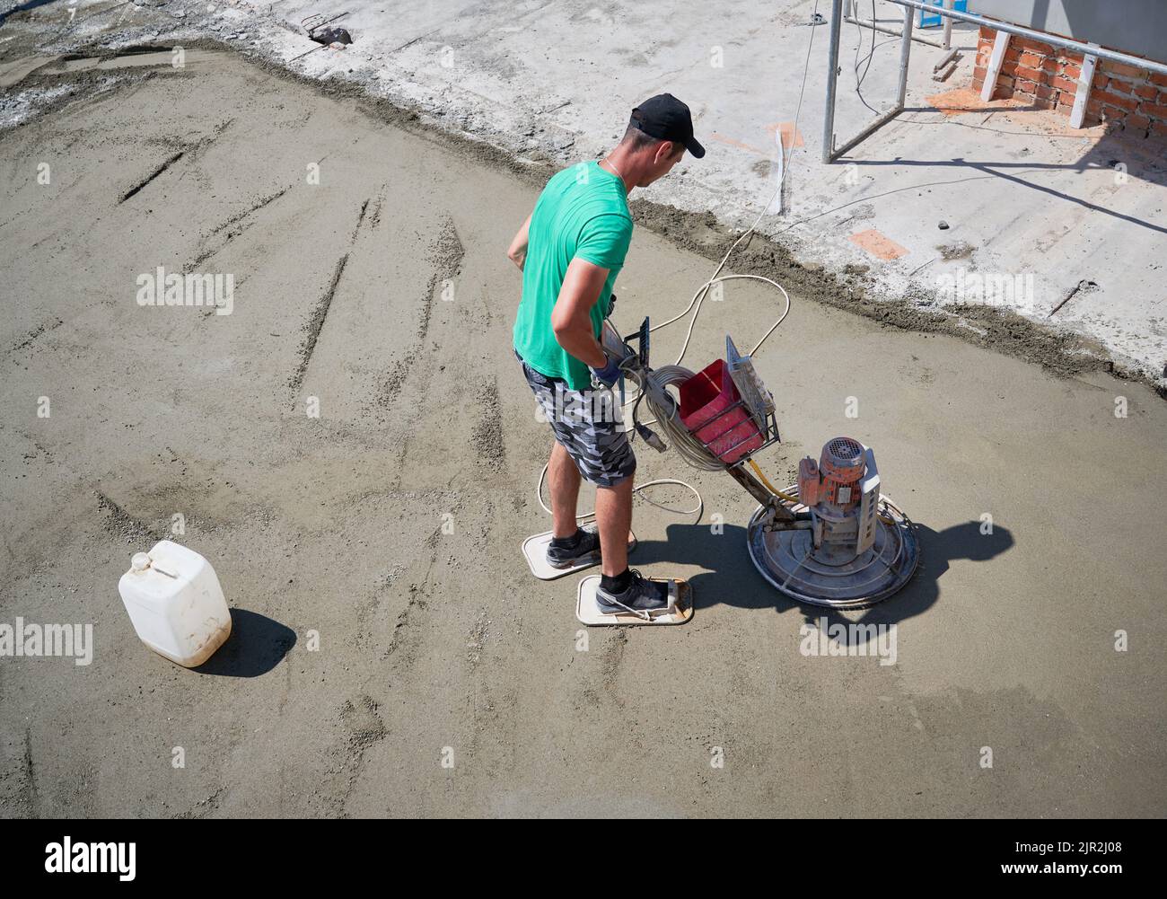 Per tutta la lunghezza di un lavoratore maschio che utilizza una macchina da trogelatura durante la spianatura del pavimento in un nuovo edificio. Uomo in pantaloncini per la finitura di superfici in calcestruzzo con smerigliatrice per pavimenti in cantiere. Foto Stock