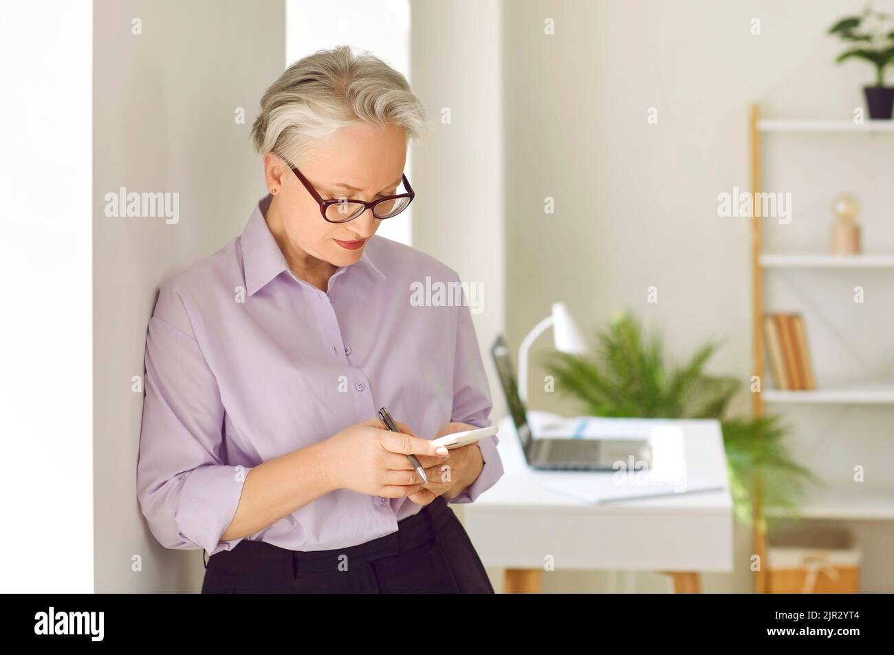 Bella donna d'affari senior in piedi in ufficio e utilizzando il suo telefono cellulare Foto Stock