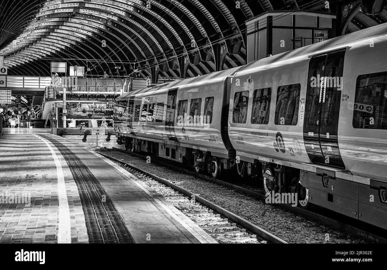 Accanto alla piattaforma della stazione ferroviaria si trova un treno. Ci sono passeggeri sullo sfondo e alcuni camminano attraverso un ponte pedonale. Una storica tettoia curva è Foto Stock