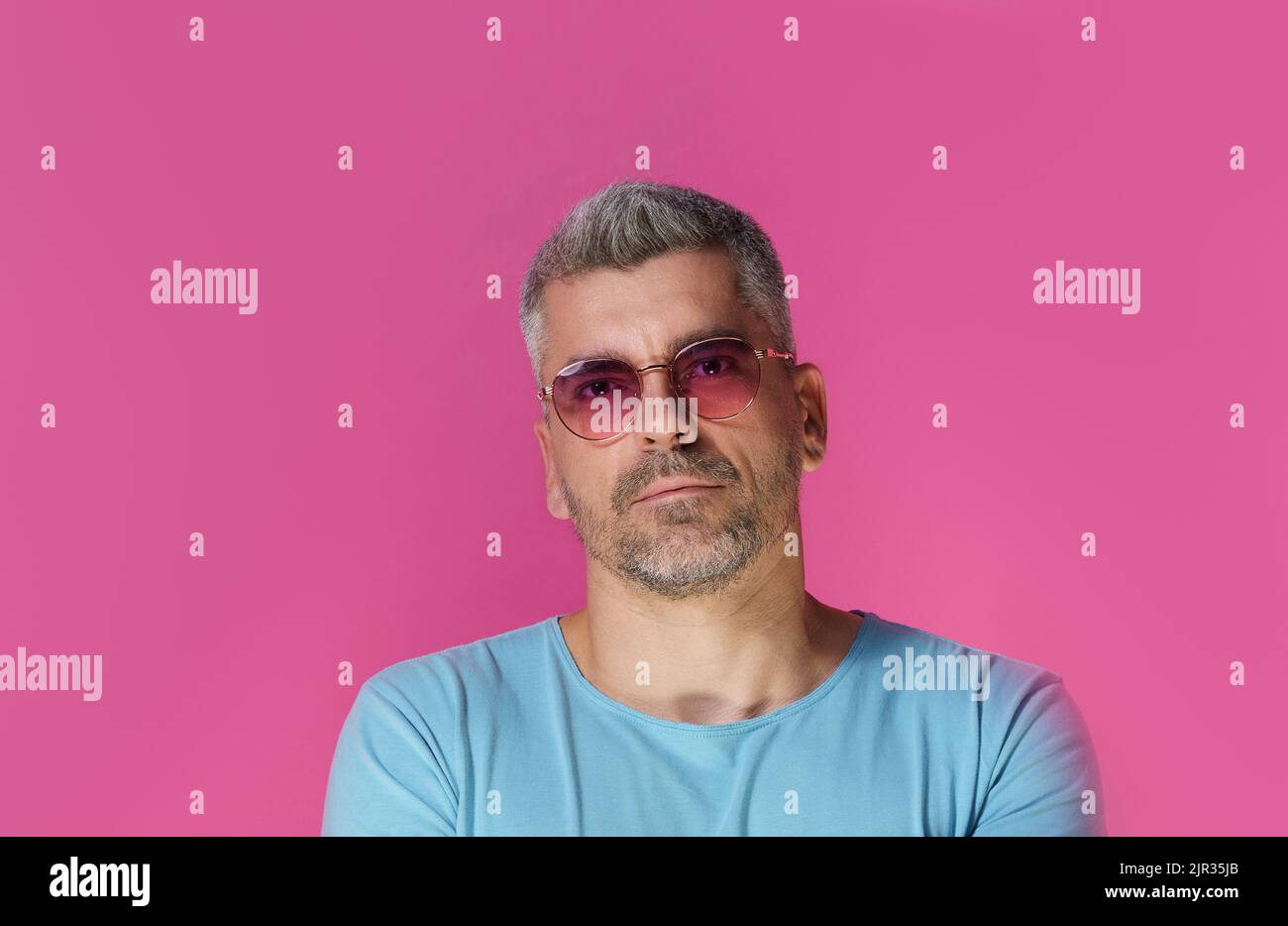 Bell'uomo 30s 40s che indossa occhiali da sole rosa e t-shirt blu con un look aggressivo e concentrato isolato su sfondo rosa. Un uomo maturo dai capelli grigi girato in studio. Foto Stock