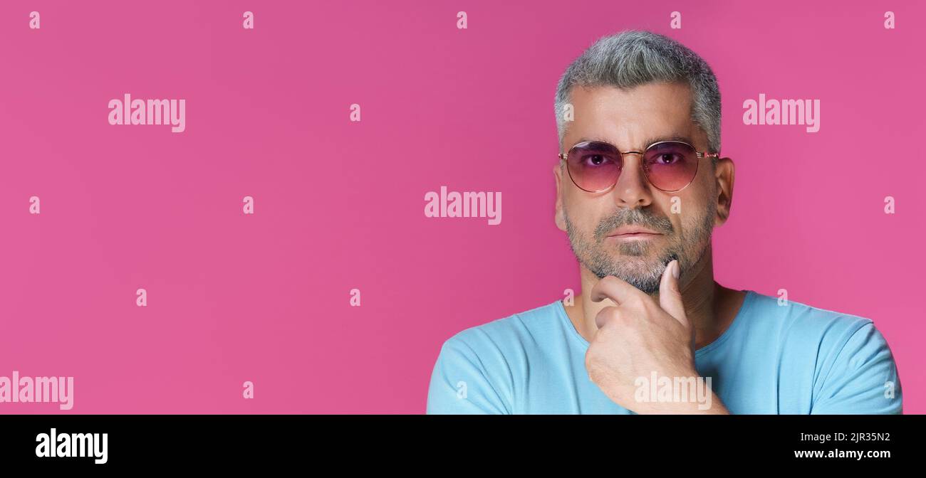 Uomo 30s 40s, molto bello, che indossa occhiali da sole rosa e una t-shirt blu che tocca il mento con un look pensoso e concentrato isolato su sfondo rosa. Un uomo maturo dai capelli grigi girato in studio. Foto Stock
