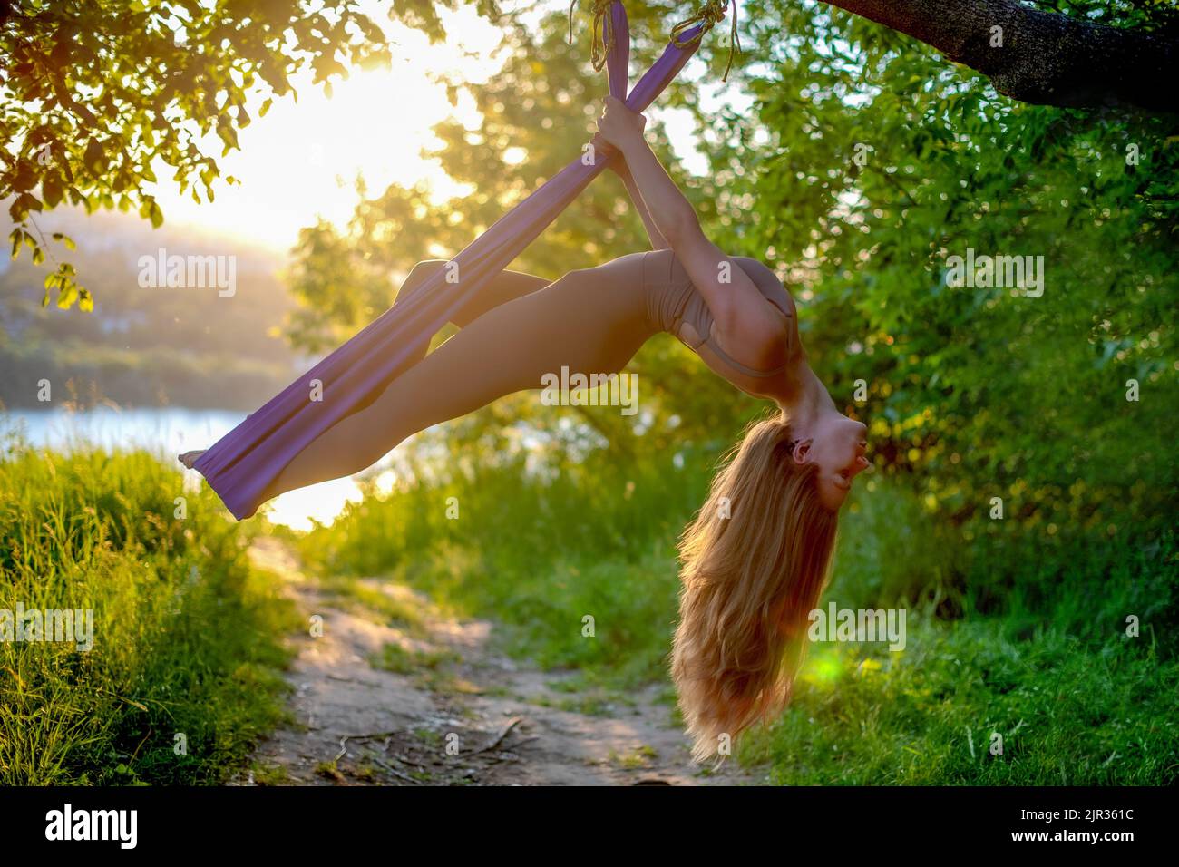 Una giovane ginnastica femminile è impegnata nello yoga aereo, utilizzando una combinazione di pose yoga tradizionali, pilates e danza utilizzando un'amaca al tramonto in natura Foto Stock