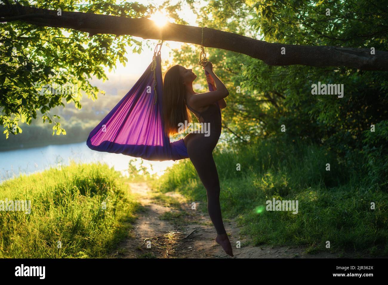 Una giovane ginnastica femminile è impegnata nello yoga aereo, utilizzando una combinazione di pose yoga tradizionali, pilates e danza utilizzando un'amaca al tramonto in natura Foto Stock