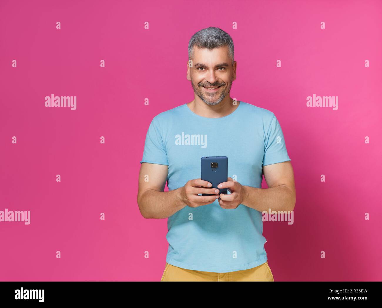 Allegro giovane uomo adulto 30s 40s in casual vestire tenere smartphone lettura di testo o la navigazione online isolato su sfondo rosa. Uomo mucho in t-shirt blu con scatto da studio per telefono. Foto Stock