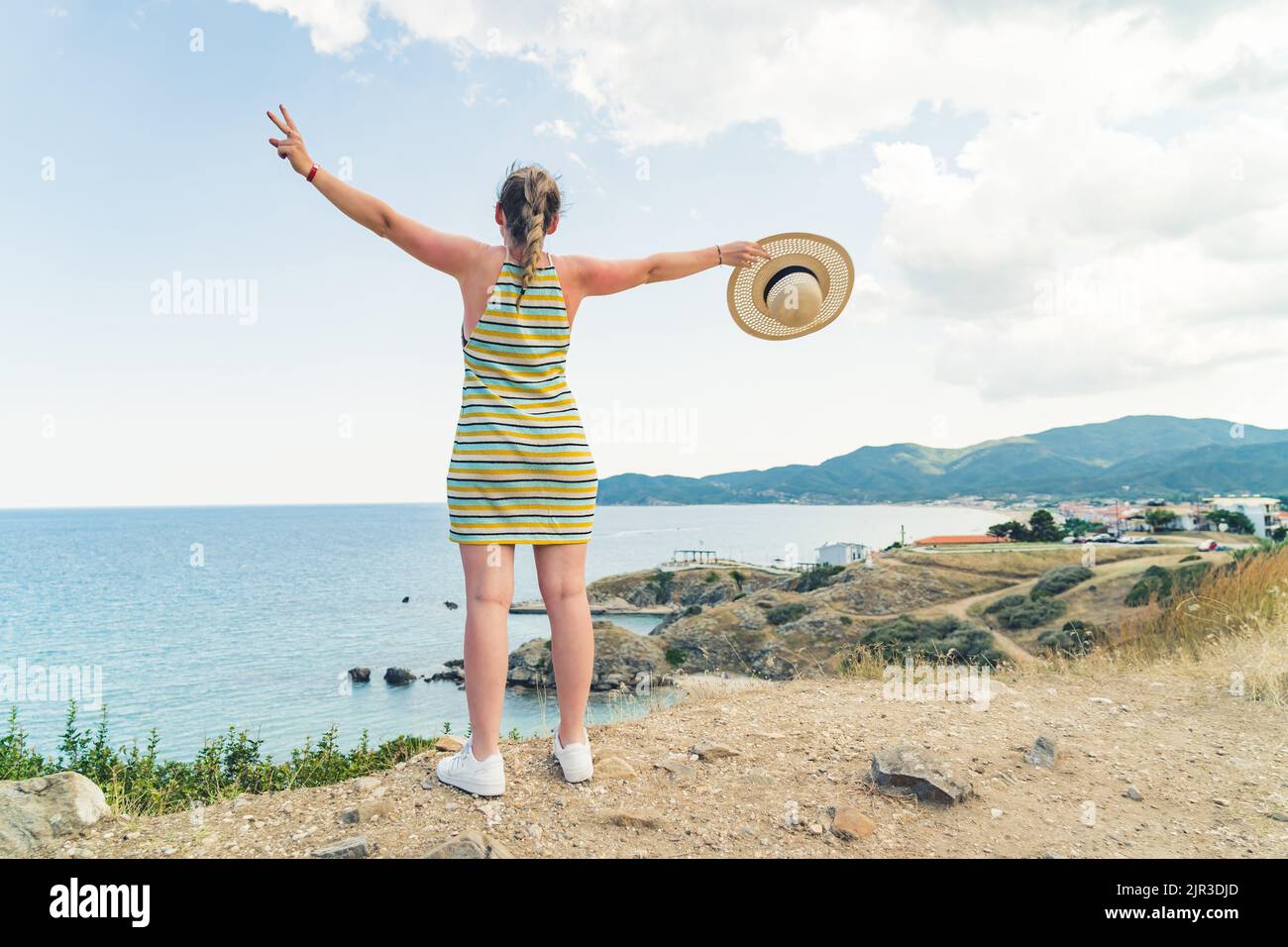 Concetto di viaggio all'estero. Giovane donna bionda caucasica in vestito a righe e sneakers bianche guardando mozzafiato greco Seascape spalancando le braccia in entrambe le direzioni. Foto di alta qualità Foto Stock