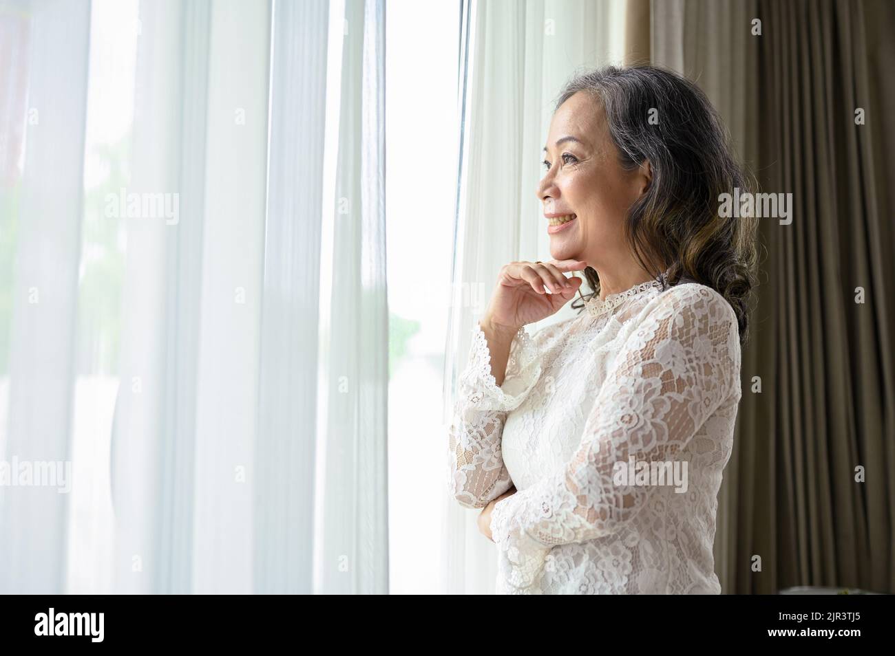 Donna asiatica di mezza età rilassata e rilassata guarda fuori dalla finestra, sognando ad occhi aperti la felicità nella sua vita. Foto Stock