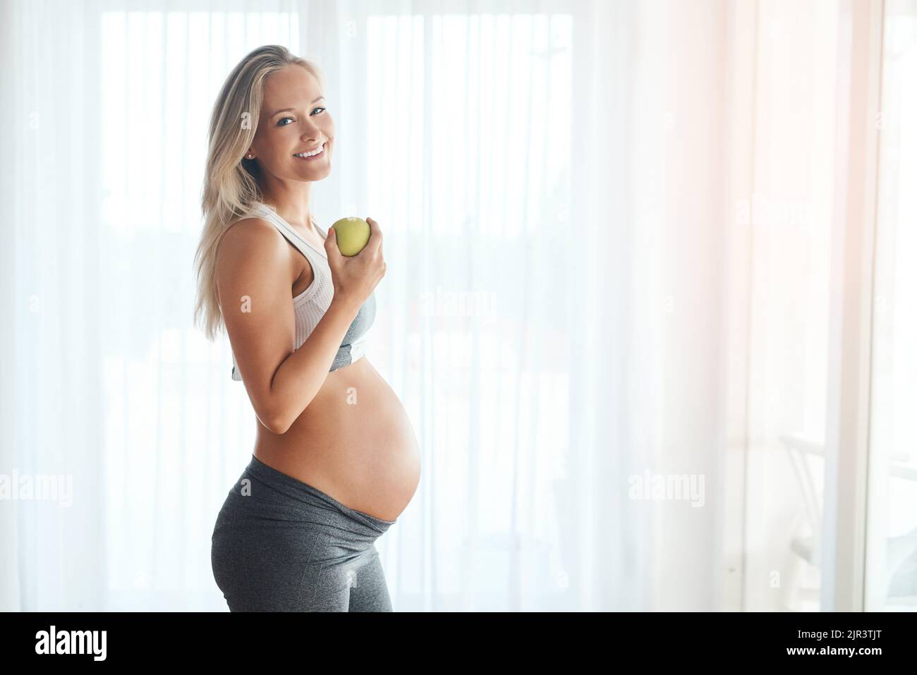 Il mio bambino lo ama quando mangio bene. Ritratto di una donna felice incinta vestita di abbigliamento sportivo e mangiare una mela a casa. Foto Stock