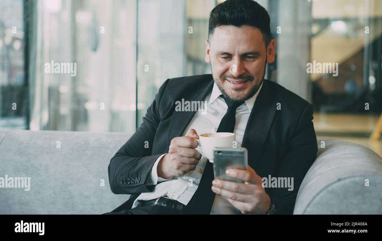 Sorridente uomo d'affari caucasico in abiti formali sorridente, rilassante e guardando lo schermo del suo smartphone mentre beve caffè in un arioso caffè durante l'ora di pranzo Foto Stock