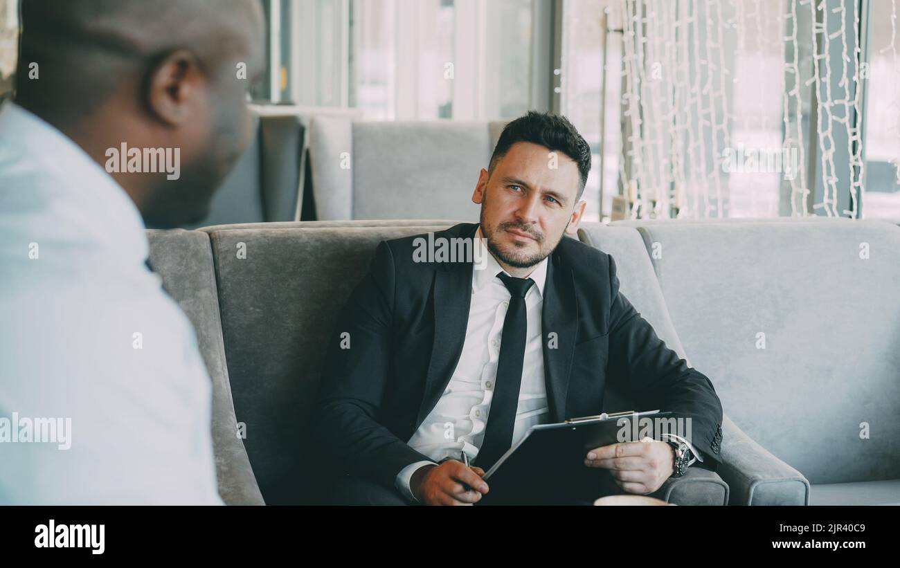 Uomo d'affari di HR che ha intervista di lavoro con l'uomo afroamericano e che guarda la sua domanda del resume in caffè moderno all'interno Foto Stock