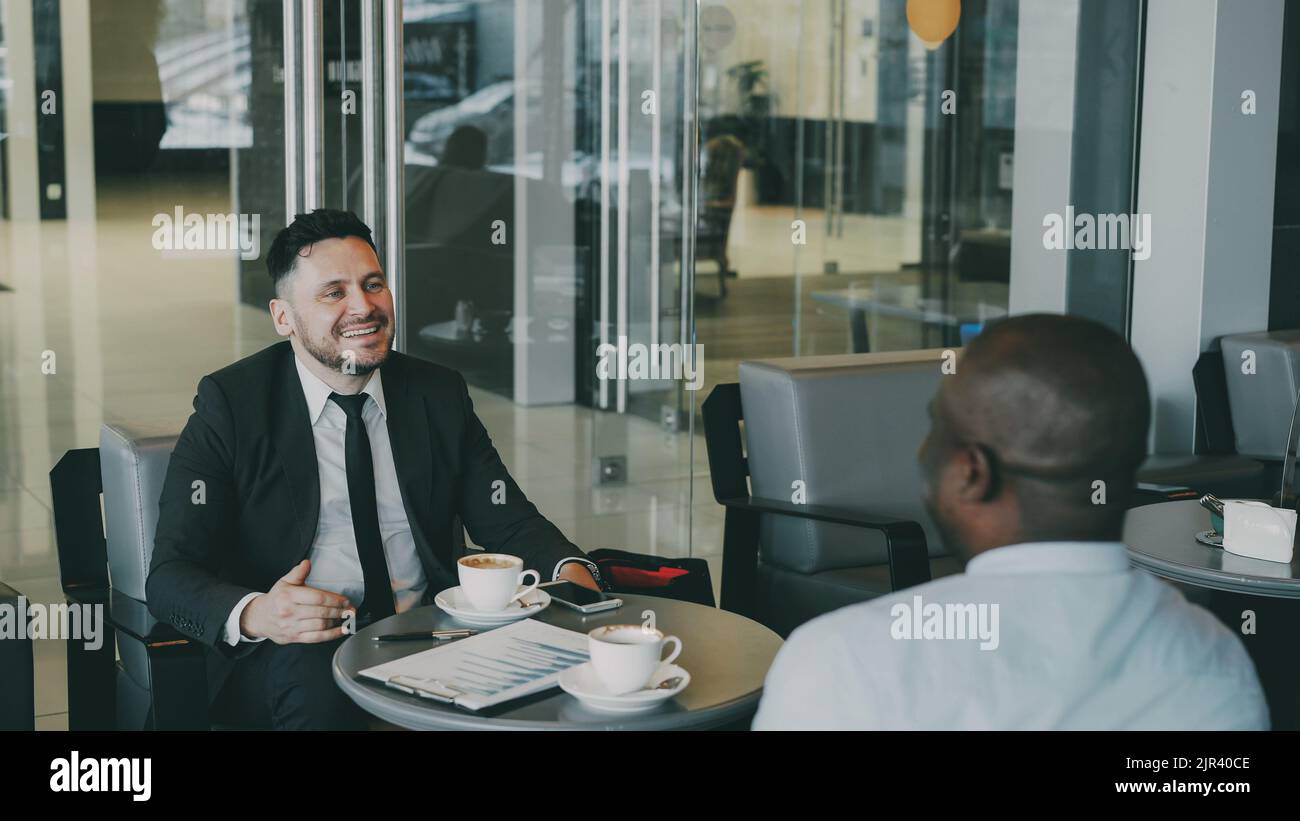 Allegro uomo d'affari caucasico bearded in abiti formali sorridendo, gesticulating, bevendo caffè e discutendo il rapporto finanziario di startup con il suo socio afro-americano in caffè moderno Foto Stock