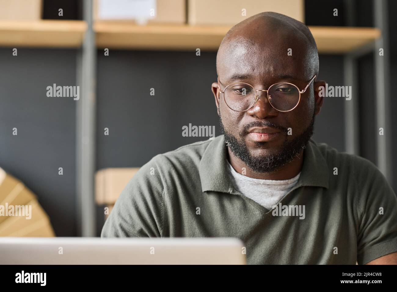 Uomo serio africano in occhiali che lavorano online utilizzando il laptop in magazzino Foto Stock