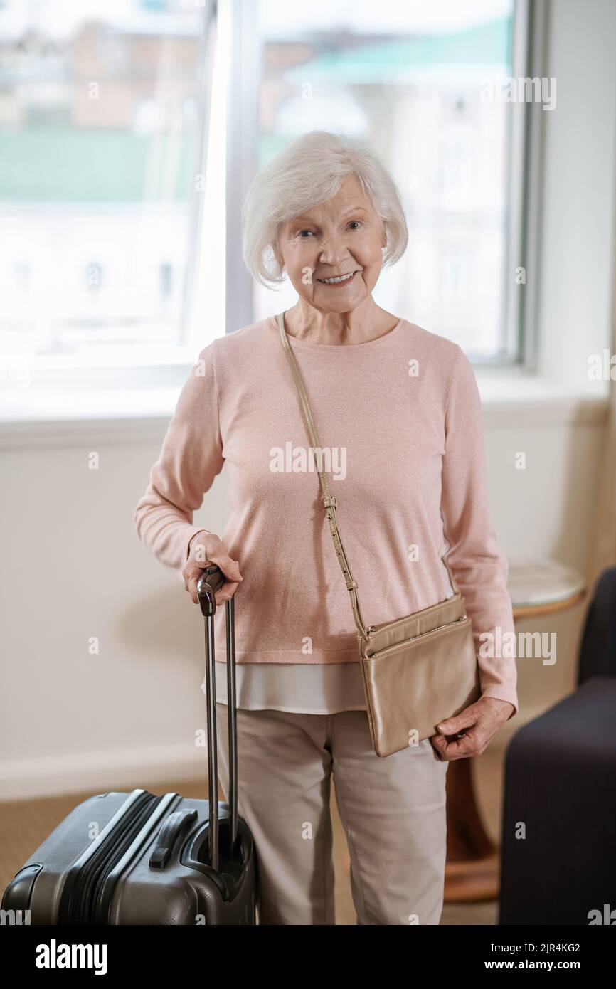 Donna anziana dall'aspetto buono in piedi con una valigia in una camera d'albergo Foto Stock