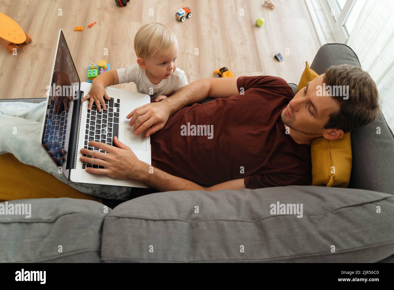 Naughty piccolo bambino che si preoccupa il padre sdraiato sul divano con il laptop nel soggiorno-stanza, che tocca la tastiera con la mano, che disturba il lavoro da casa o che riposa il papà. Figlio toddler che chiede l'attenzione del papà Foto Stock