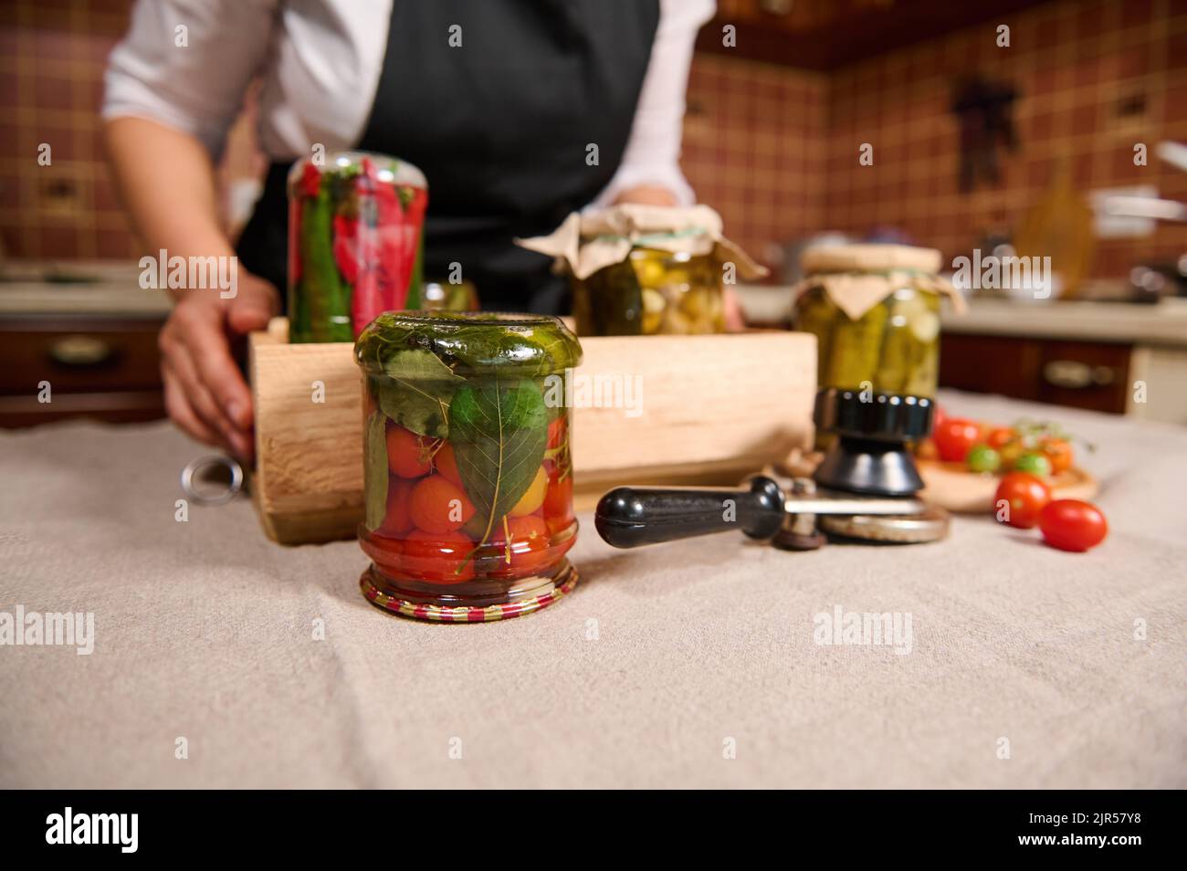 Concentrati su vasetto di pomodori ciliegini marinati, su un tavolo vicino a una chiave di aggraffatura e una scatola di verdure in scatola. Inscatolamento. Decapaggio Foto Stock