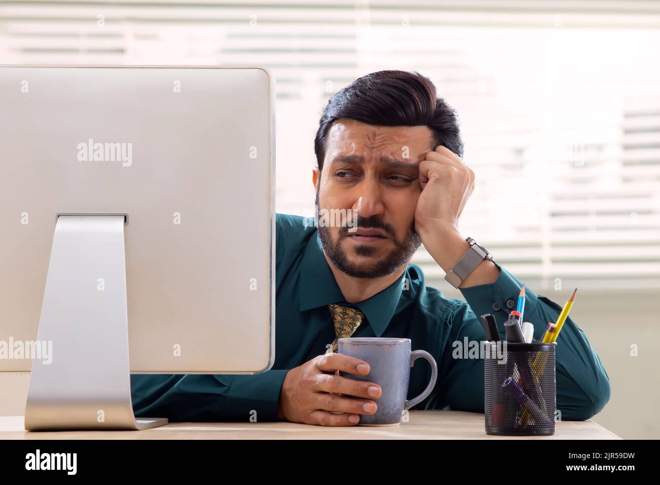 Un dipendente aziendale in abbigliamento formale che guarda con disinteresse lo schermo del desktop con occhi sonnolenti in ufficio. Foto Stock