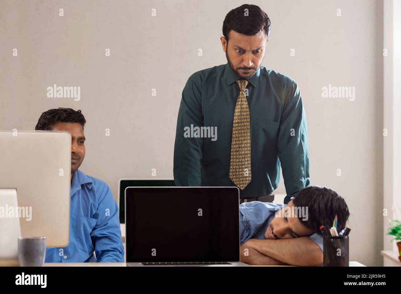 Un boss arrabbiato dietro due dipendenti aziendali, uno dei quali dorme alla scrivania. Foto Stock