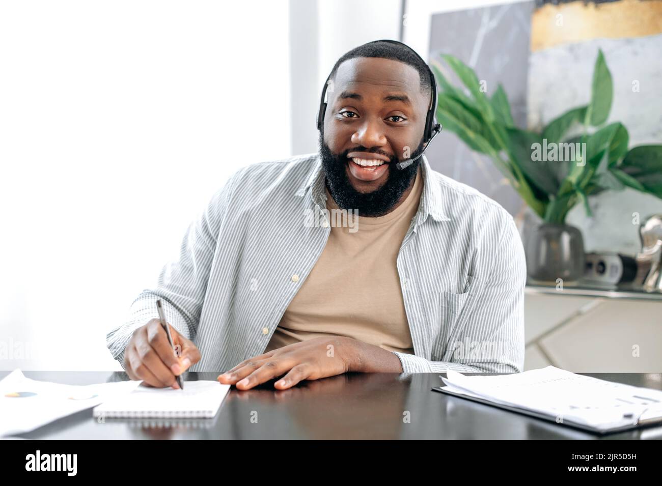 Uomo afro-americano fiducioso con cuffie, leader aziendale, dipendente aziendale, avere una conversazione online, ascolta l'interlocutore, conduce la consultazione, prende appunti, sorride Foto Stock