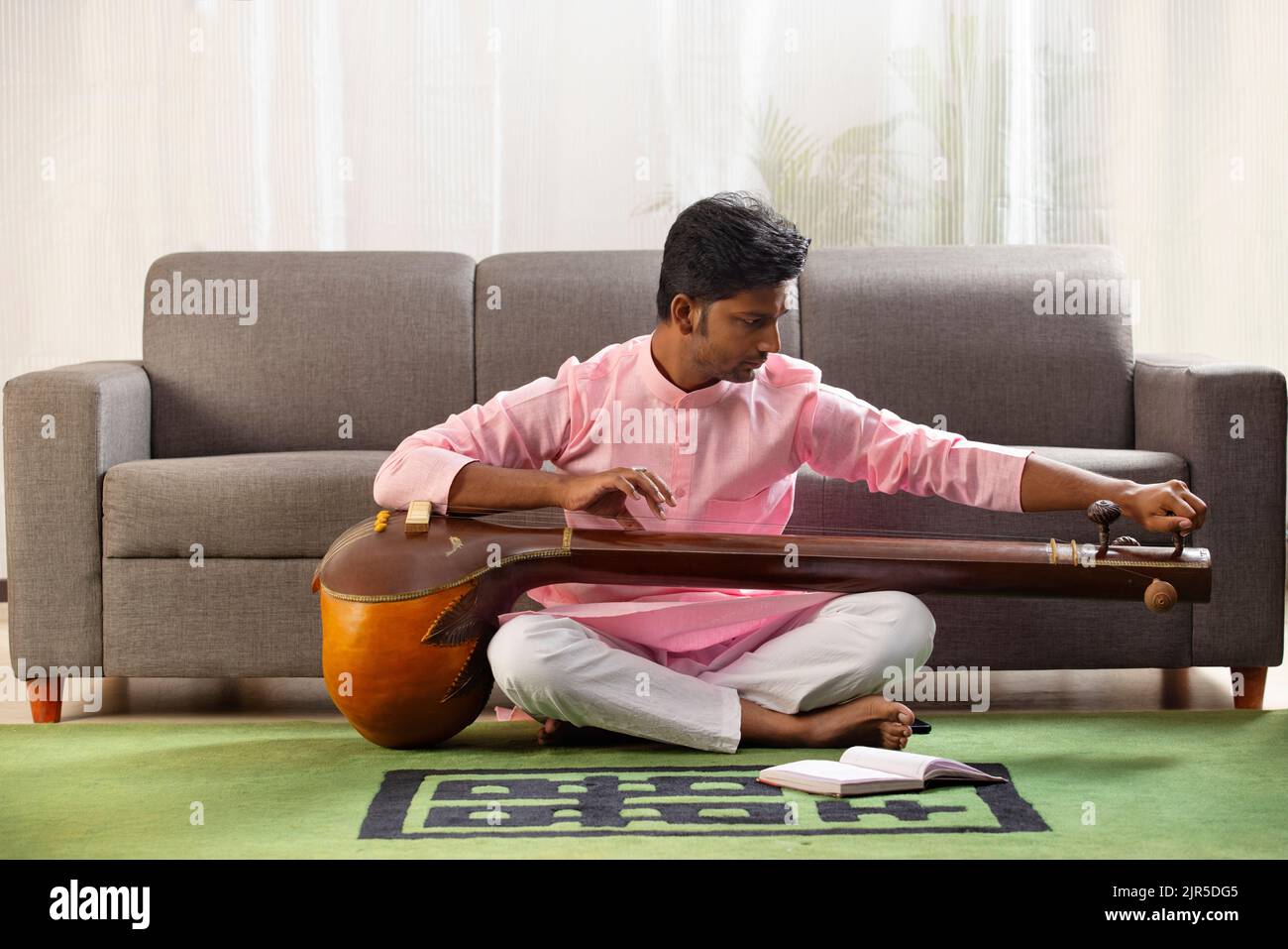 Giovane musicista maschio che sintonizza il suo tanpura mentre si siede in salotto Foto Stock