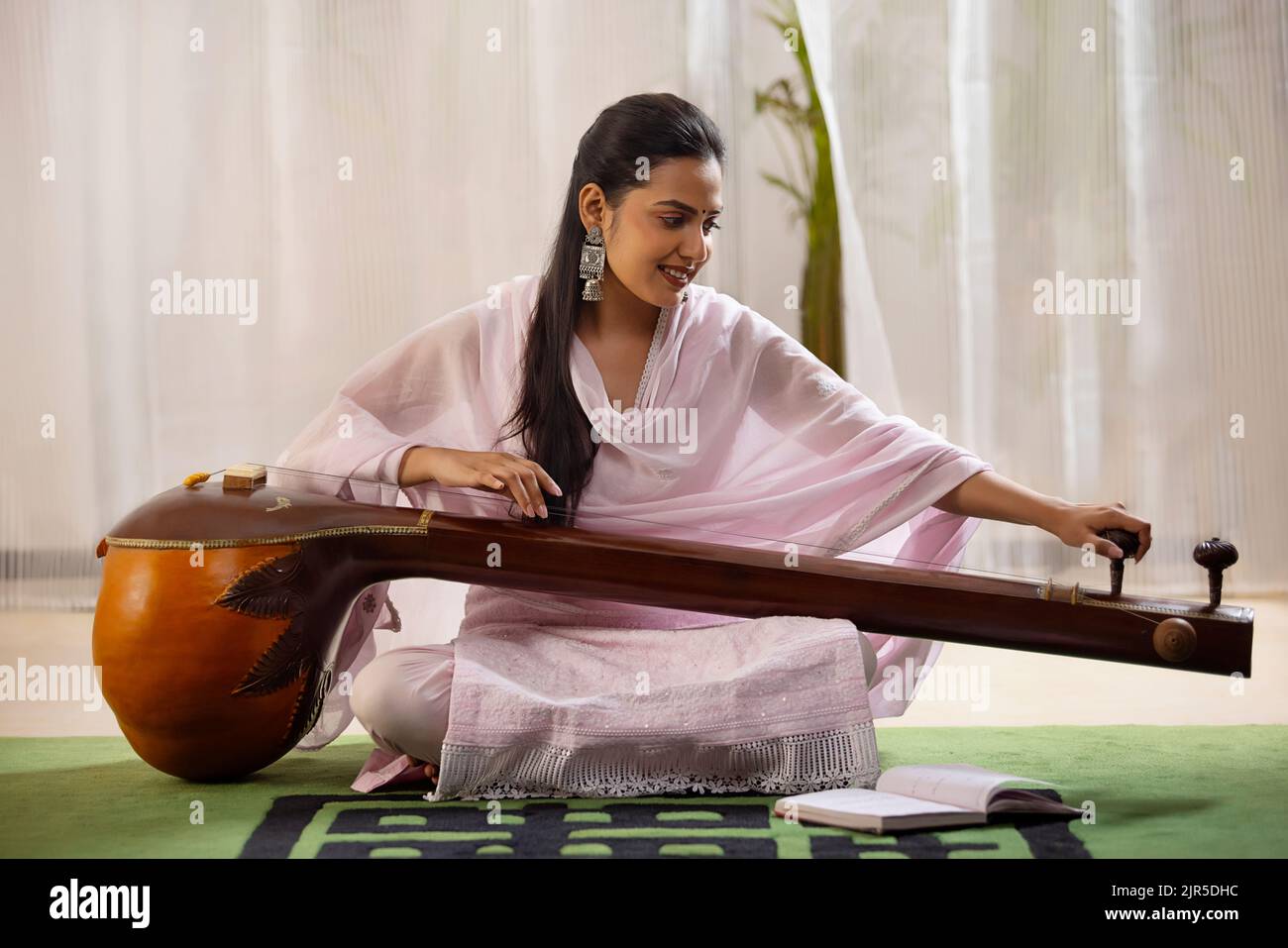 Giovane musicista femminile che sintonizza la sua tanpura mentre si siede in salotto Foto Stock