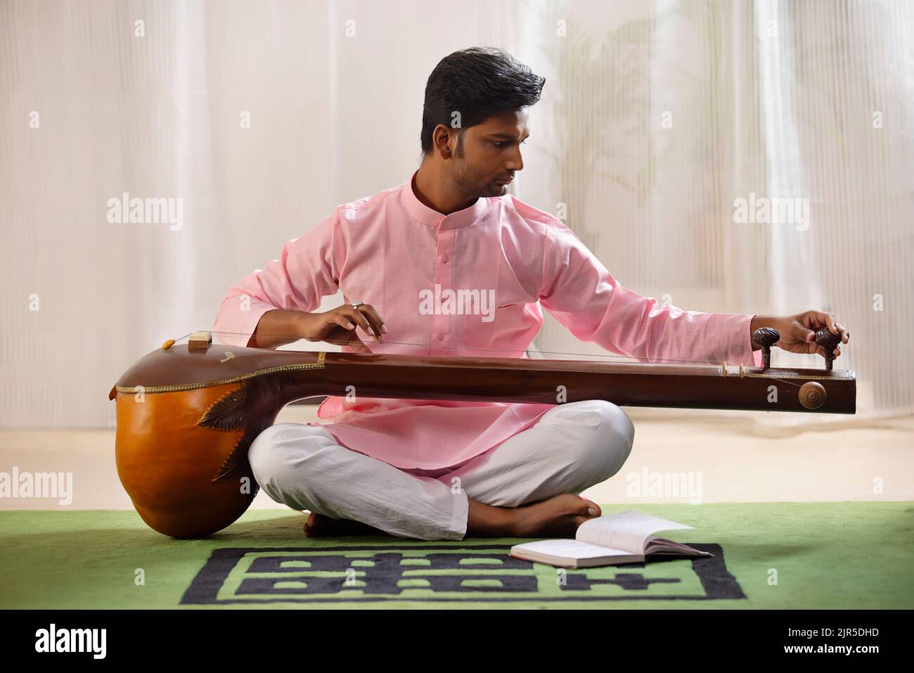 Giovane musicista maschio che sintonizza il suo tanpura mentre si siede in salotto Foto Stock