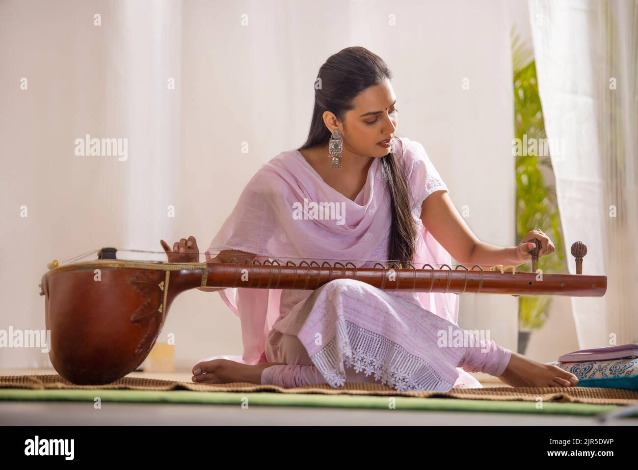 Giovane musicista femminile che sintonizza il suo sitar mentre si siede nel salotto Foto Stock