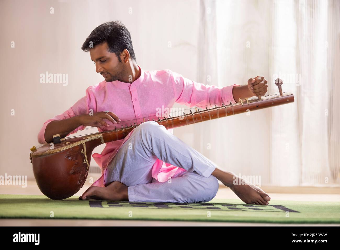 Giovane musicista maschio che sintonizza il suo sitar mentre si siede nel soggiorno Foto Stock