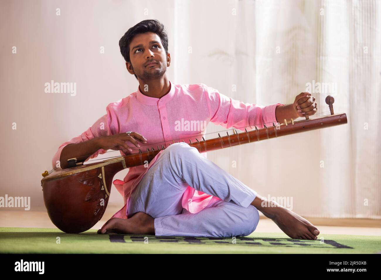 Giovane musicista maschio che sintonizza il suo sitar mentre si siede nel soggiorno Foto Stock