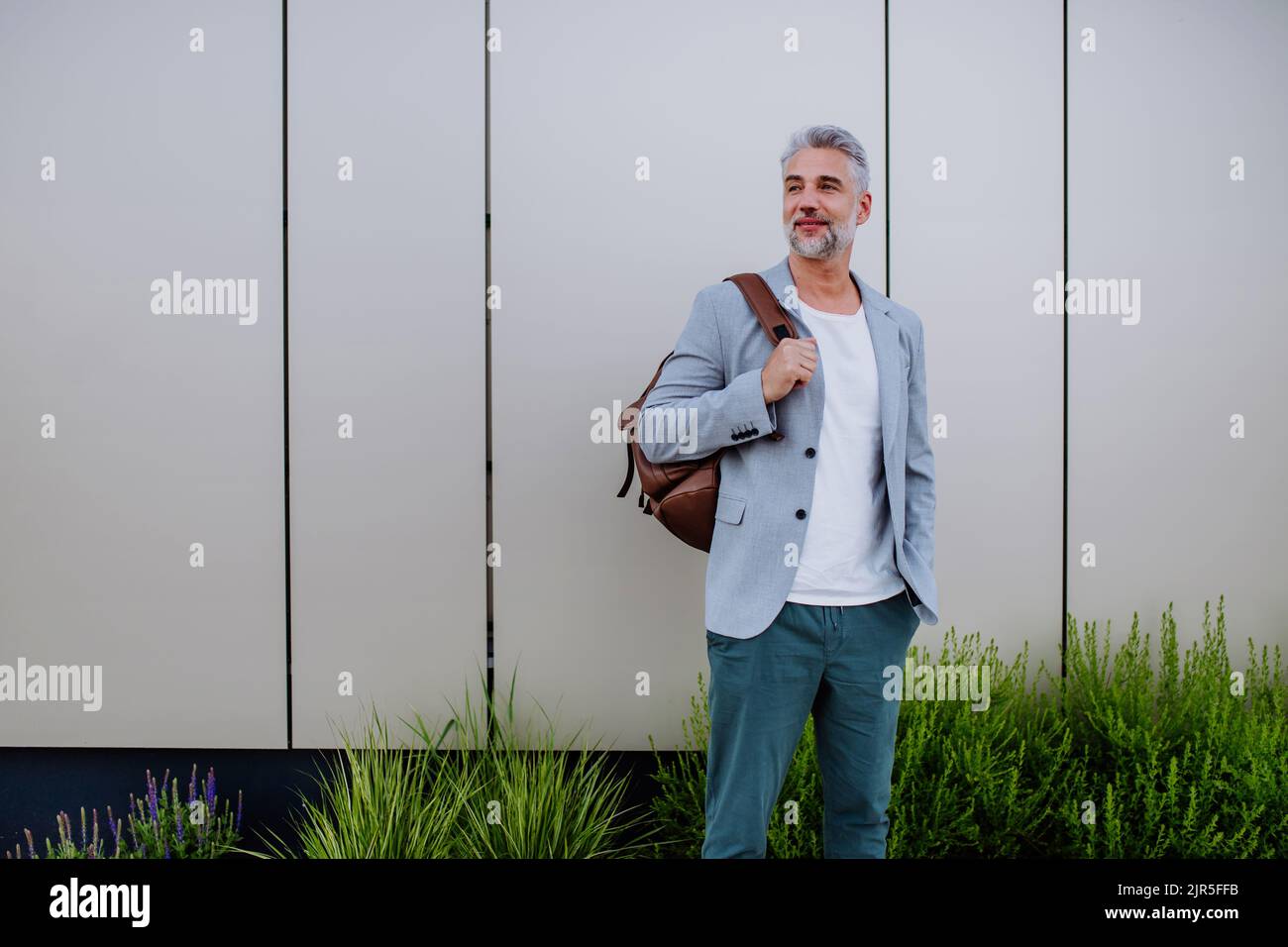 Uomo d'affari maturo e rilassato con zaino appoggiato sulla parete della strada, concetto di equilibrio della vita lavorativa. Foto Stock