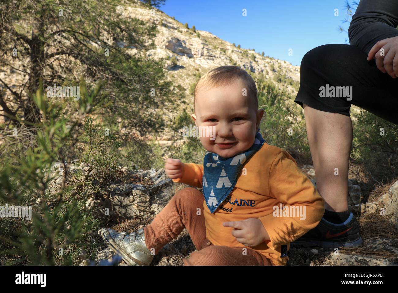 Bambino caucasico seduto nella foresta giocando con una roccia e sorridendo in direzione della macchina fotografica Foto Stock
