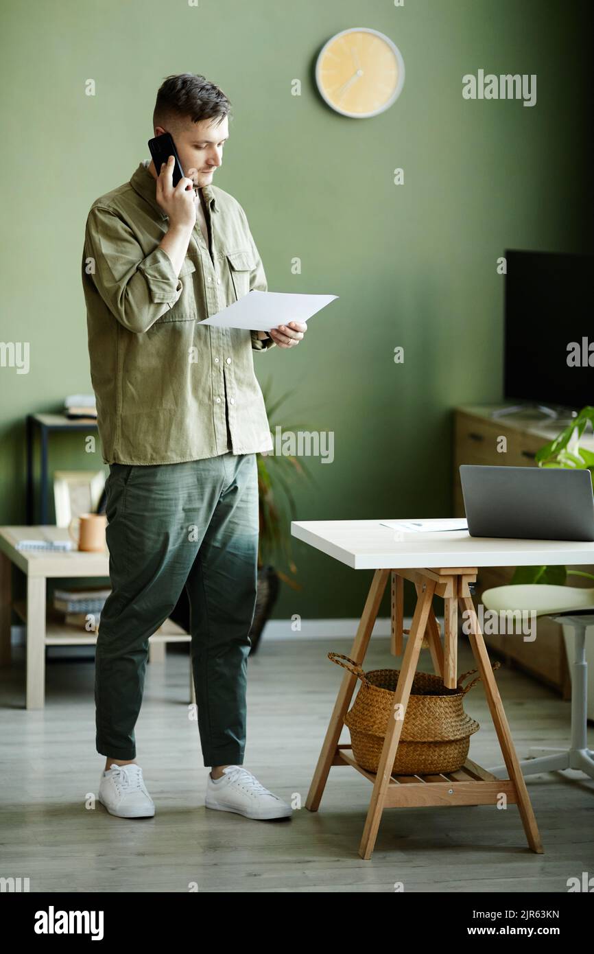 Giovane uomo che discute di un documento aziendale durante una conversazione sul telefono cellulare mentre si è in piedi nella stanza Foto Stock