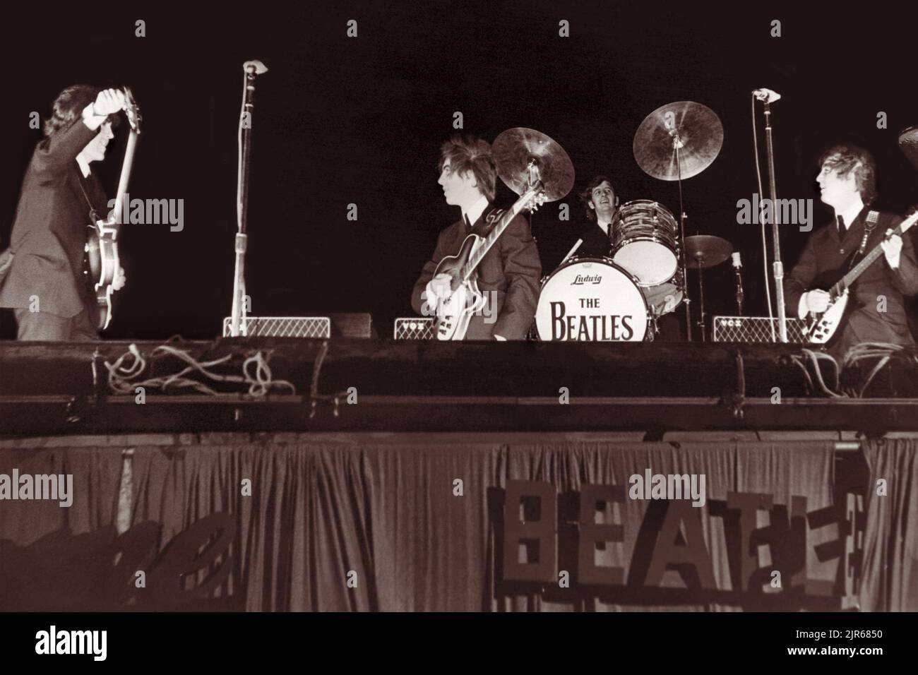 I Beatles in scena al Gator Bowl di Jacksonville, Florida, il 11 settembre 1964. L'uragano Dora aveva colpito Jacksonville il giorno precedente e i venti forti rimasti, con una raffica di 45 miglia all'ora, richiesero che la batteria di Ringo Starr fosse inchiodata sul palco e che la scritta "Beatles" di cartone sul lato del palco fosse finalmente strappata via. (USA) Foto Stock