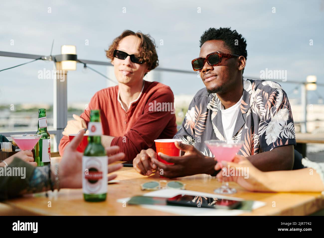 Due ragazzi interculturali in occhiali da sole e casualwear sorseggiando un drink nel caffè all'aperto mentre si siede al tavolo davanti ai loro amici durante la chat Foto Stock