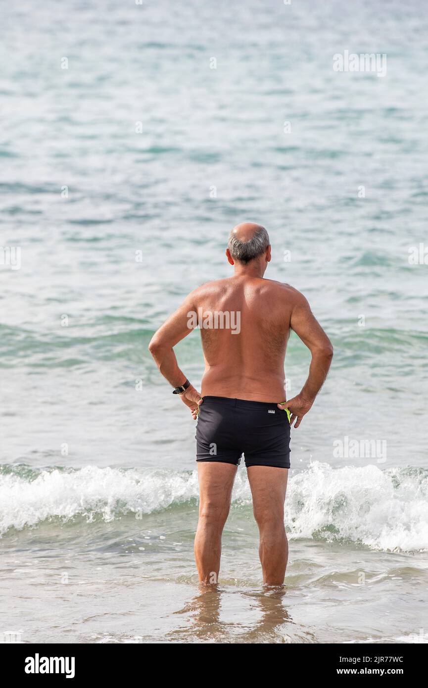 uomo che indossa costume da bagno in piedi sulla spiaggia guardando il mare, uomo più anziano sulla spiaggia che indossa piccoli costume da bagno, pensionato gentleman paddling. Foto Stock