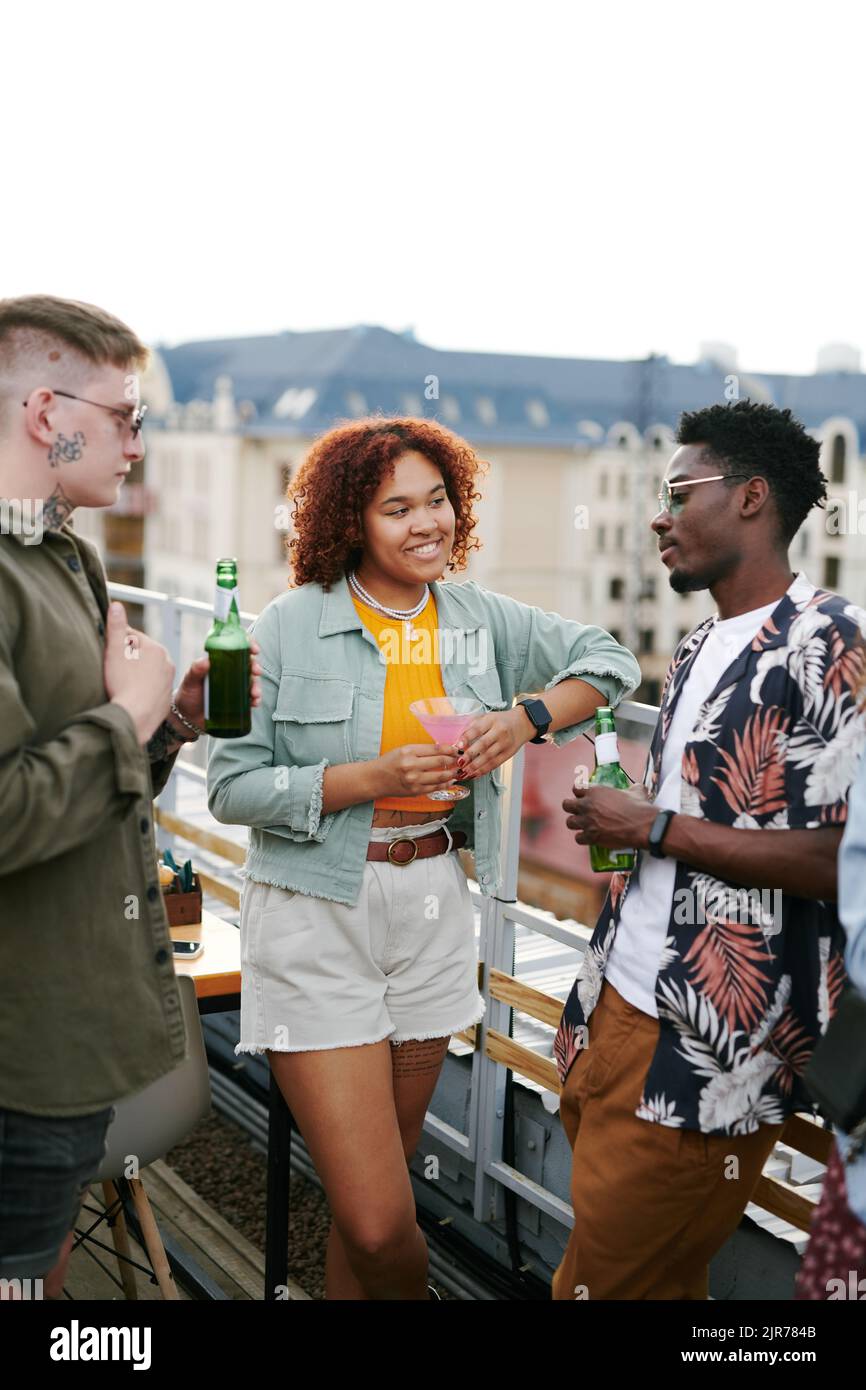 Due giovani coppie interculturali in un elegante casualwear sorseggiando un drink e chiacchierando sulla terrazza del caffè sul tetto durante una festa all'aperto Foto Stock
