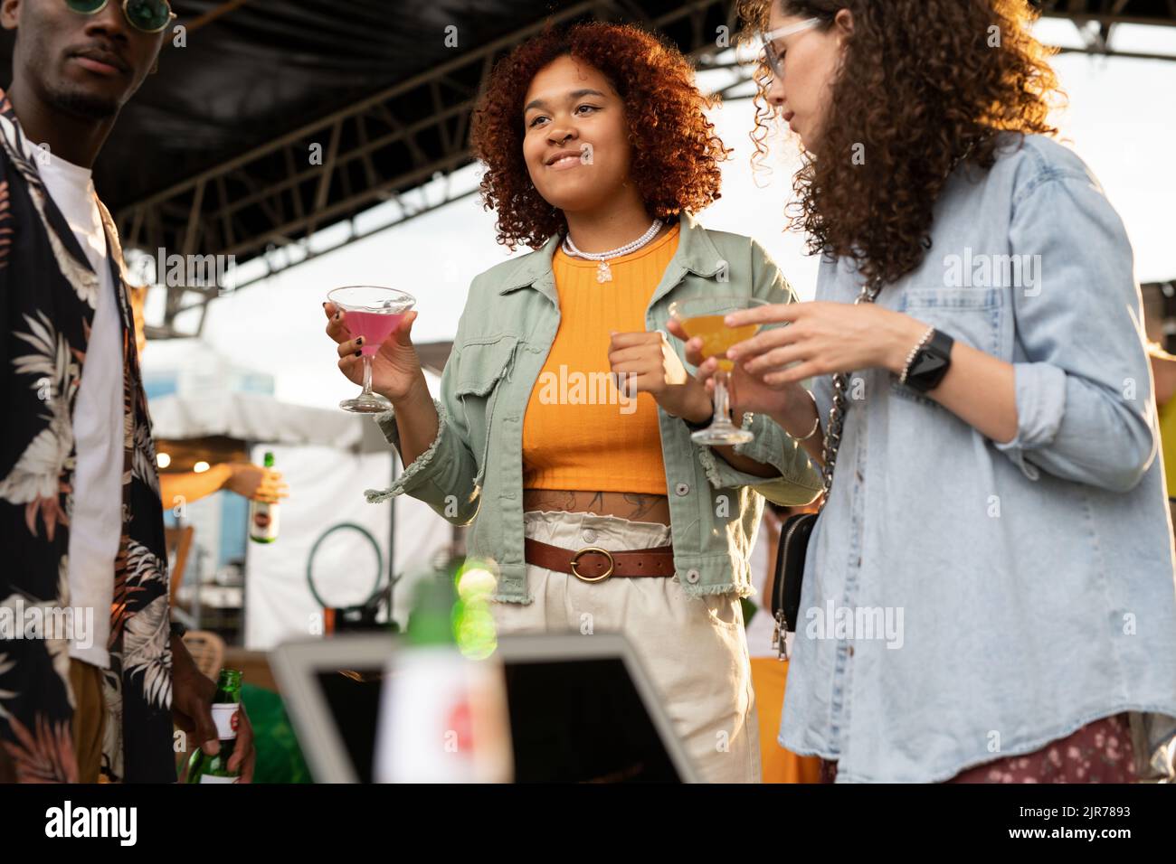 Due ragazze interculturali con cocktail e un ragazzo afroamericano che si gode la festa sul tetto mentre si trova in piedi in un caffè all'aperto nel fine settimana estivo Foto Stock