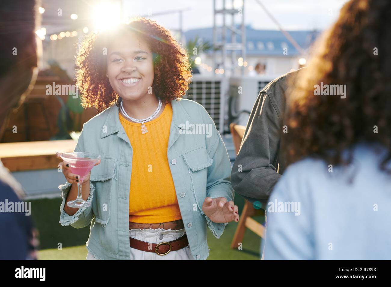 Felice giovane donna con un cocktail guardando uno dei suoi amici mentre si gode la festa sul tetto o la riunione in caffè all'aperto Foto Stock