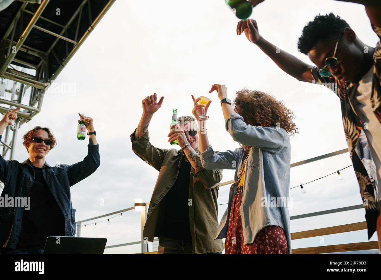 Gruppo di giovani amici estatici con bottiglie di birra che ballano al party sul tetto in un caffè all'aperto mentre si è in piedi con le braccia sollevate Foto Stock