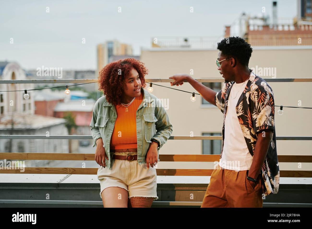 Giovane coppia afro-americana in elegante casualwear chiacchierando sulla terrazza del caffè sul tetto contro l'ambiente urbano Foto Stock