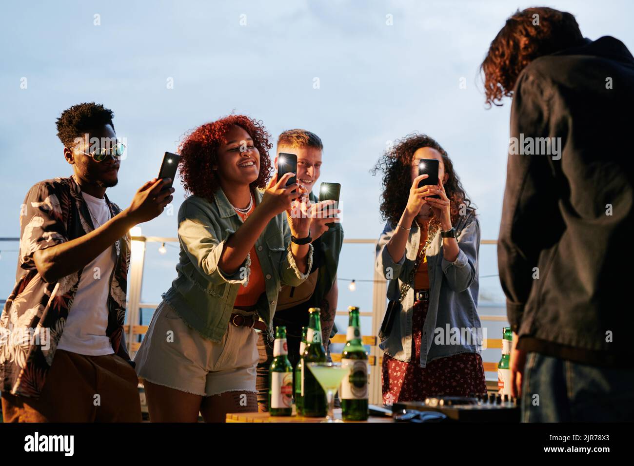 Due giovani e felici coppie interculturali con smartphone che scattano foto di deejay al party sul tetto mentre si trovano di fronte a lui Foto Stock