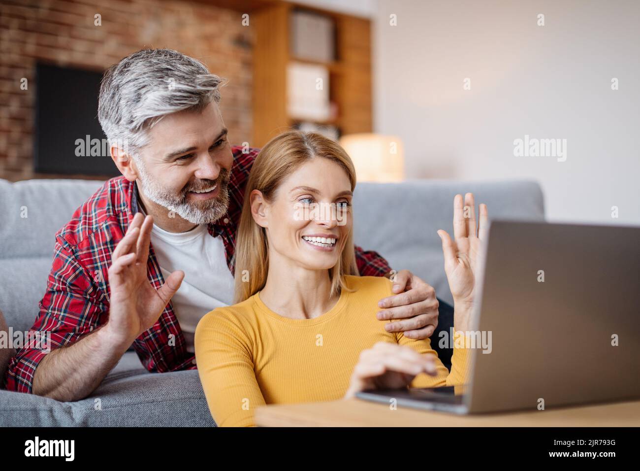 Felice sorridente marito caucasico di mezza età abbracciando moglie, facendo una mano nel laptop, facendo una chiamata in linea Foto Stock