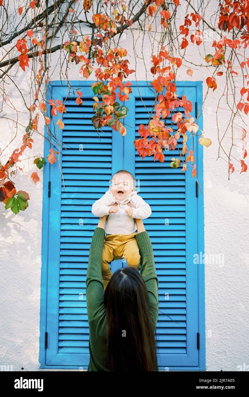 La mamma tiene un bambino ridente alto sopra la sua testa vicino ai ciechi sulla finestra Foto Stock