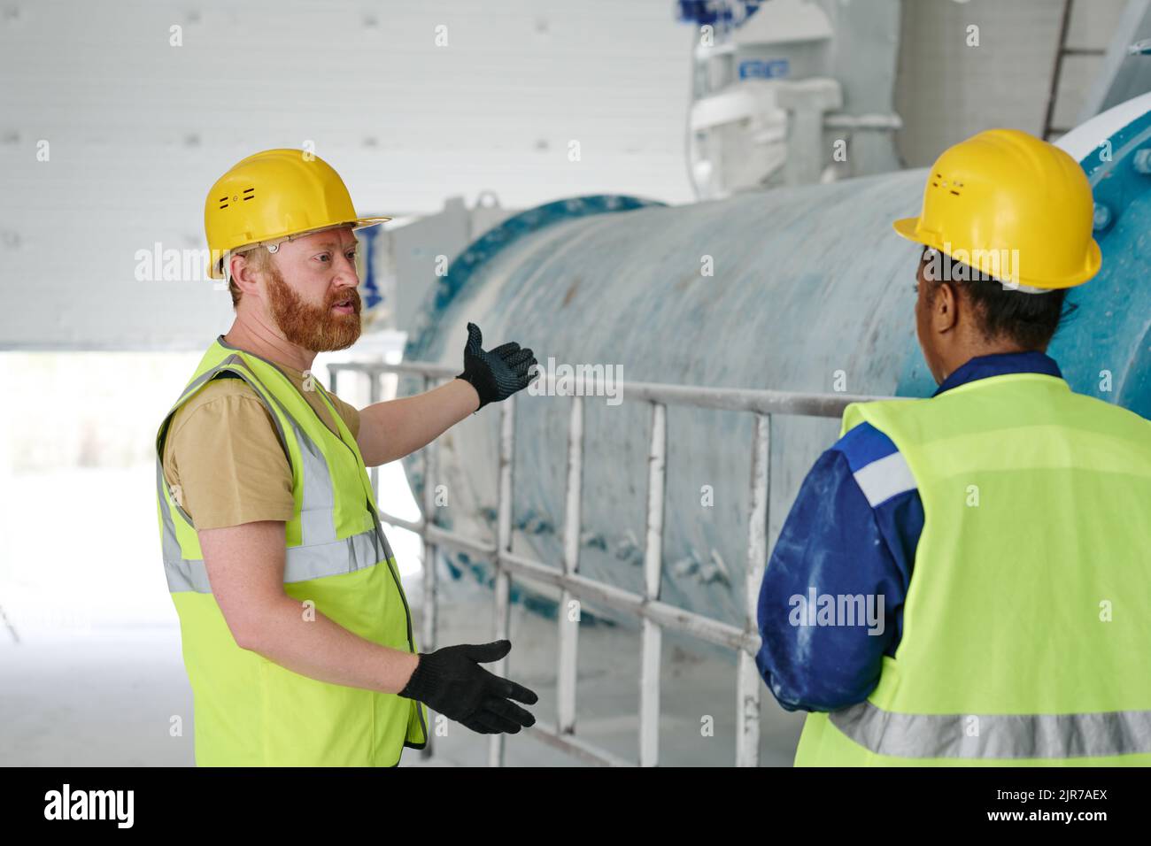 Uomo sicuro ingegnere in uniforme, guanti e casco di sicurezza parlare con una giovane collega afro-americana durante il lavoro in fabbrica Foto Stock