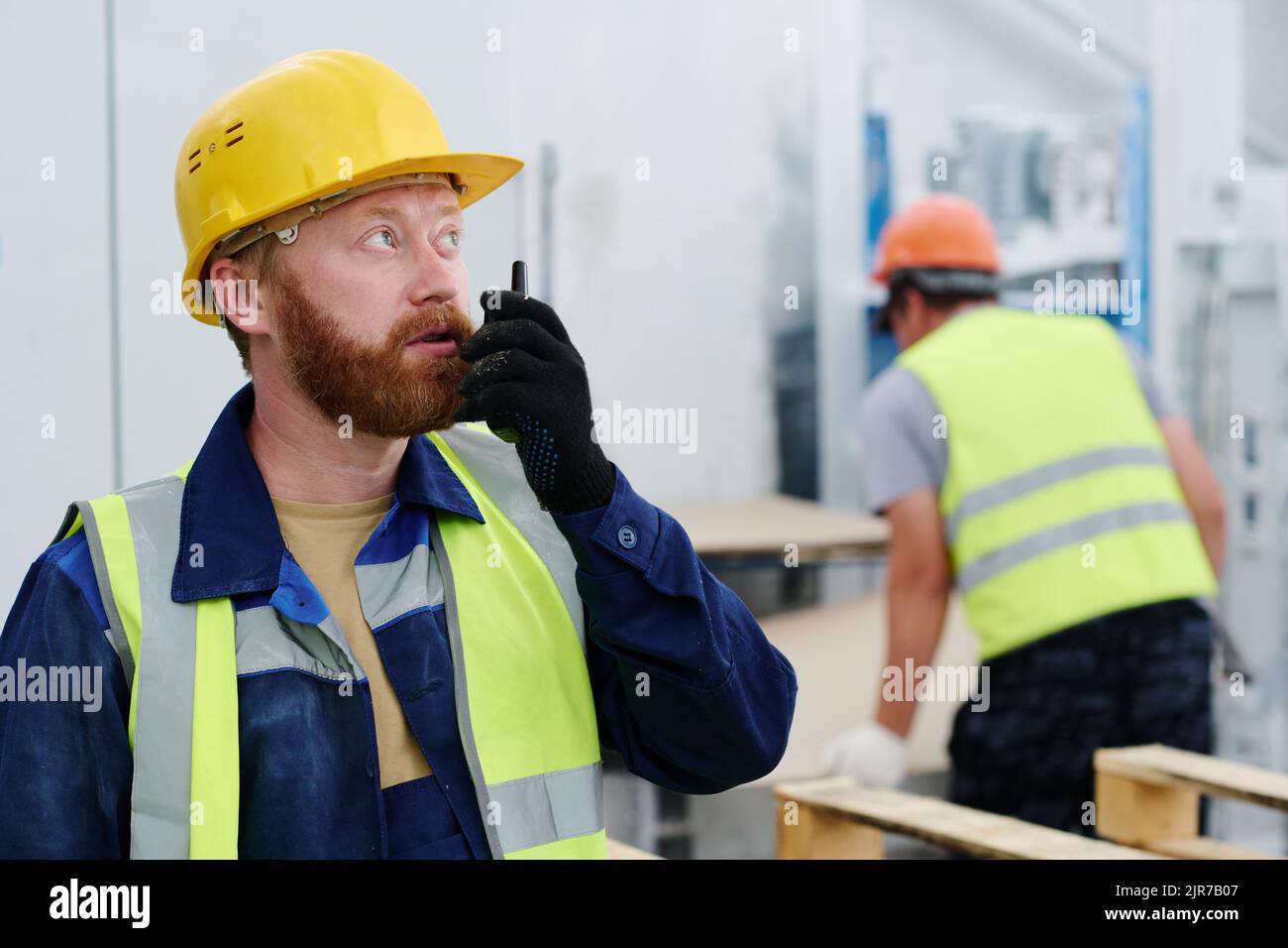 Giovane lavoratore fiducioso di fabbrica che parla in walkie-talkie mentre si trova davanti alla telecamera contro il collega in officina Foto Stock