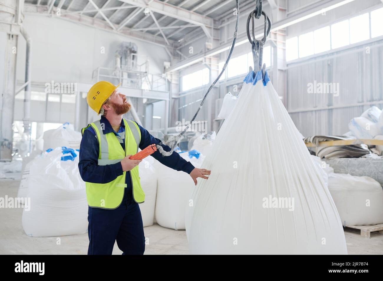 Giovane ingegnere maschile che guarda verso l'alto un enorme gancio dove sacco bianco con la materia prima appeso mentre si controlla il processo di sollevamento Foto Stock