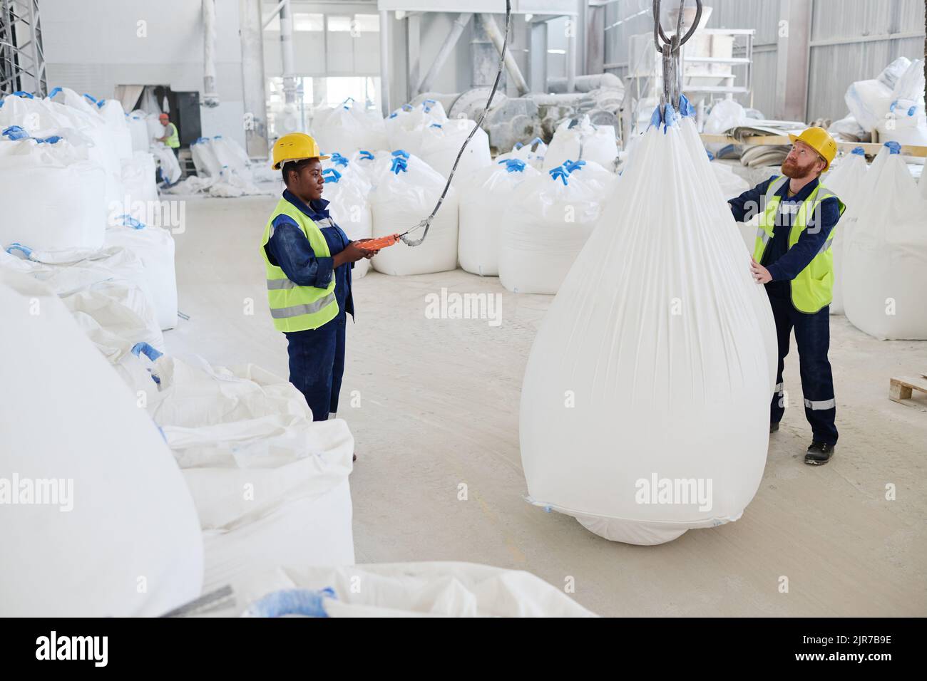 Un giovane lavoratore maschile che aiuta la sua collega con l'interruttore di comando a distanza a sollevare enormi sacchi pesanti con materie prime in officina Foto Stock