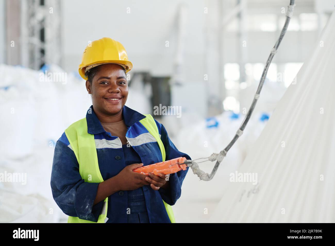 Felice giovane ingegnere di fabbrica utilizzando il telecomando interruttore mentre sollevano enormi sacchi bianchi pesanti e guardando la fotocamera Foto Stock